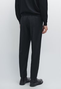 Person iført sorte skræddersyede bukser, sorte Chelsea-støvler og en sort langærmet top, stående med ryggen til mod en ensfarvet baggrund.