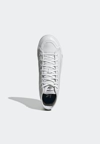 White canvas sneaker with a rounded toe, lace-up design, and rubber outsole. Features a black and white logo patch on the tongue.