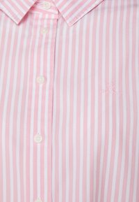 Růžovo-bíle pruhovaná košile s knoflíkovým zapínáním, špičatým límcem a malým vyšitým logem dole. Hladká bavlněná textura.