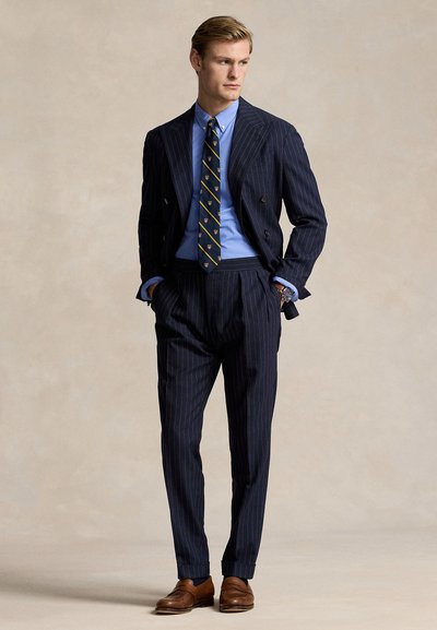 Férfi tengerészkék vékony csíkos dupla breasted öltönyben, kék inggel, mintás nyakkendővel, barna bőr mokaszinokkal, zsebében a kezeivel, jobbra néz.