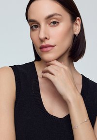 Czarny dzianinowy top bez rękawów z szerokim dekoltem, na nadgarstku srebrna bransoletka z okrągłymi detalami. Gładki odcień skóry i neutralne tło.