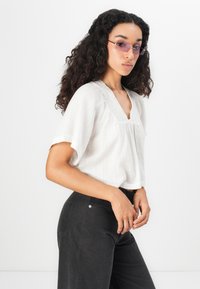 Vit blus med kort ärm i texturerad tyg och broderad halsringning, matchad med högmidjade svarta byxor och lila solglasögon.
