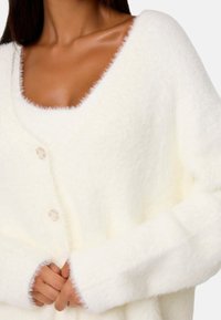Cardigan bianco morbido con scollo a V profondo, tre bottoni e maniche lunghe. La texture sfumata e il materiale soffice creano un aspetto accogliente.