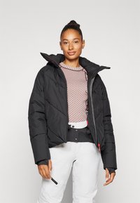 Doudoune noire avec capuche, présentant un design matelassé, des poches zippées et des accents rouges, portée par-dessus un pull à motif rose clair et un pantalon blanc.