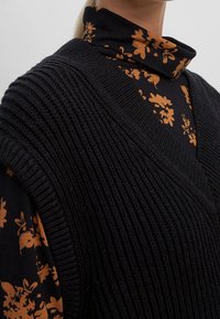 Maglione nero lavorato a maglia con scollo a V profondo, indossato sopra un top nero con motivo floreale arancione e colletto alto. Superficie strutturata visibile.