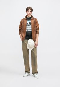 Brązowy dziergany kardigan na czarnej koszulce z nadrukiem, spodnie khaki, białe trampki i kremowa czapka, stojący na neutralnym tle.