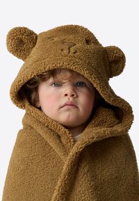 Pruun karupoeg-teemaline kapuutsiga tekk, valmistatud pehmest tekstureeritud kangast, kõrvadega ja tikitud näoga, lapsel ümber rippumas.