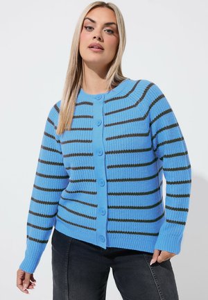 Woman with long blond hair wears a blue knit cardigan with black horizontal stripes and black pants against a plain light background.