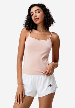 Frau mit lockigem Haar, die ein hellrosa Top und weiße Sportshorts mit schwarzem Logo trägt, posiert vor einem schlichten Hintergrund.