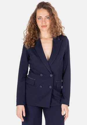 Donna con capelli ricci che indossa una giacca doppiopetto blu navy e pantaloni abbinati, in piedi davanti a uno sfondo bianco uniforme.