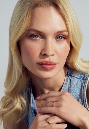 Person med ljust hår iført en denimvest, med glat hud. Guld ringe på fingrene, med en neutral baggrund. Blå øjne og neutral makeup.