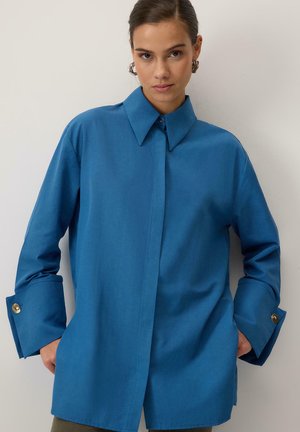 Jauna sieviete zilā pārmērīgi lielā kreklā ar platām manžetēm un metāla pogām, stāvot pret vienkāršu gaišu fonu.