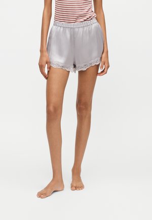 Femme portant un short en satin gris clair avec bordure en dentelle et une chemise rayée rouge et blanche, debout pieds nus sur un fond blanc.