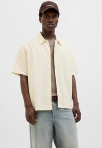 Camisa de manga corta, texturizada, de color crema con un cierre de botones, llevada sobre una camiseta de color beige claro. Combinada con jeans de color azul claro y una gorra marrón.
