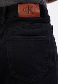 Poche arrière d’un jean en denim noir avec un patch logo Calvin Klein en cuir marron sur la ceinture.