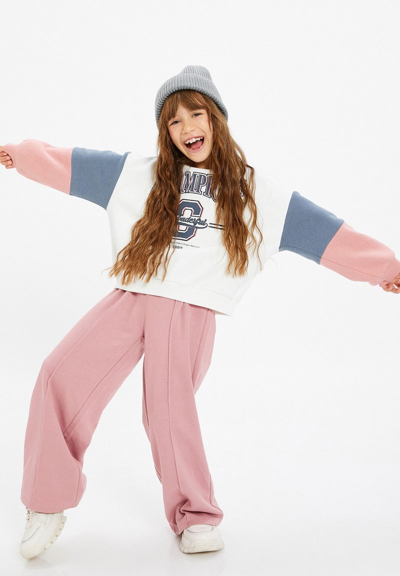 Witte sweatshirt met roze en blauwe mouwen, voorzien van een logo. Draagt het met lichtroze wijde broek en witte sneakers. Grijze beanie op het hoofd.