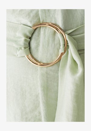 Tissu vert clair avec une finition douce et texturée. Présente un grand anneau circulaire doré mettant en valeur un détail noué.