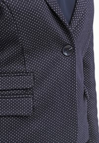 Marinblazer med vita prickar, enkel knappstängning, slag i kragen och sidofickor. Strukturert tyg tillför visuell variation.