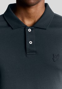 Mörkgrå polotröja av slätt tyg, med klassisk krage, två knappar och broderad logotyp på vänster sida.