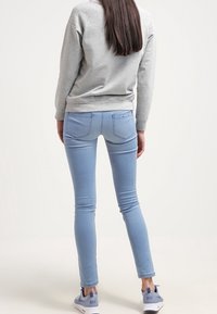 Donna in piedi, che indossa jeans skinny blu chiaro, felpa grigia e sneakers Nike grigie, vista di spalle contro uno sfondo semplice.