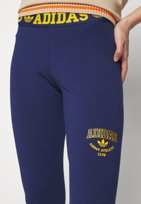 Temno modri leggingi z rumenim logotipom "ADIDAS" in besedilom "ADIDAS ATHLETIC CLUB". Imajo širok rebrast pas z oranžnimi in rumenimi črtami.