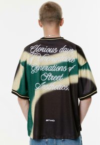 Korte mouwen, oversized shirt met een hexagonaal patroon, in groene en bruine tinten, en met witte tekst op de achterkant met de opschrift "Glorieuze dagen van Ontelbare Generaties van Straataesthetiek."