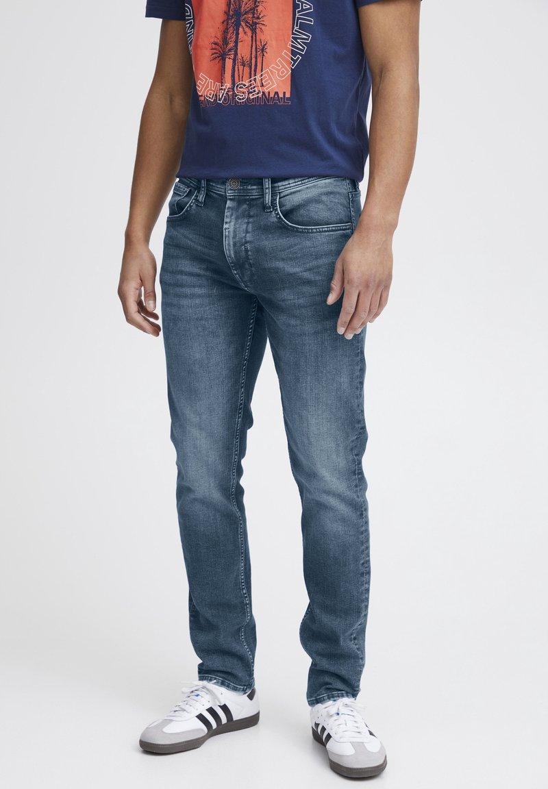 Homme portant un jean bleu délavé, un t-shirt bleu marine avec un motif de palmier orange, et des baskets blanches avec des rayures noires, posé devant un fond blanc.