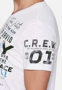 Bílá krátká tričko s krátkými rukávy, které obsahuje černý text a grafiku, včetně "C.R.E.W. 01" na rukávu a různými tištěnými prvky na přední straně.