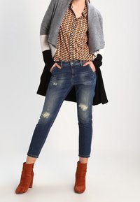 Blå denimjeans med slitna lappar, stylade med en mönstrad brun skjorta och en gråsvart cardigan. Matchade med bruna ankelboots i mocka.