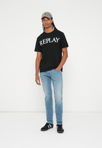 Crna majica s logotipom "REPLAY", plave uske traperice, crne cipele s bijelim prugama i siva kapa. Jednostavan dizajn i ležeran kroj.