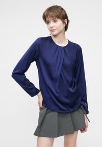 Giovane donna con capelli corti, che indossa una blusa a maniche lunghe blu navy e una gonna grigia plissettata, in piedi con una mano sulla coscia.