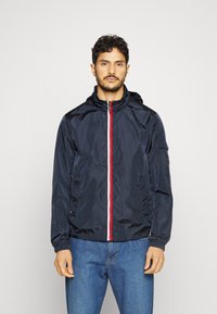 Giacca leggera della marina con zip frontale, dettagli a strisce rossa e bianca, cappuccio regolabile, polsini elastici e tasche laterali.