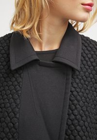 Cardigan negro texturizado con un cuello liso, que presenta un patrón tejido en forma de burbujas. El interior es de tela negra lisa, creando un efecto de capas.