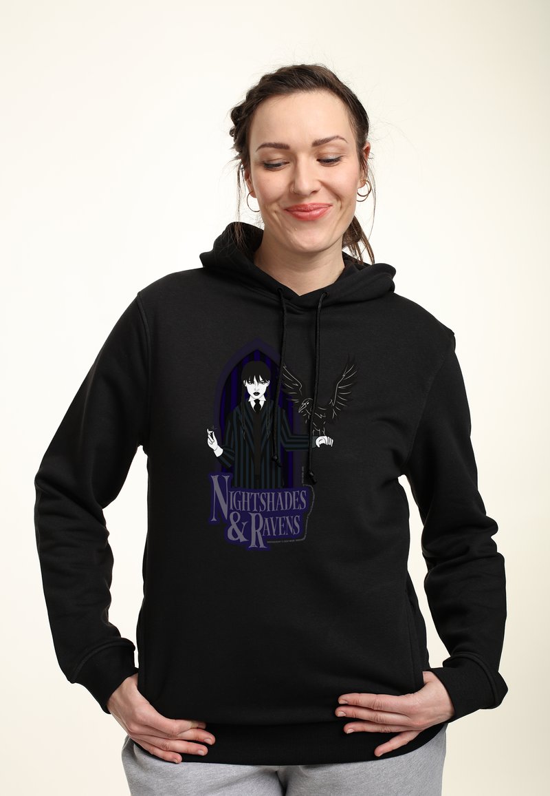 Donna che indossa un hoodie nero con la grafica di una persona in un completo a righe che tiene un corvo e il testo "Nightshades & Ravens" sulla parte anteriore.