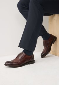 Brune lædersko med brogue-detaljer, rundet tå, snøredesign, kombineret med mørkeblå habitbukser og sokker. Træoverflade under.