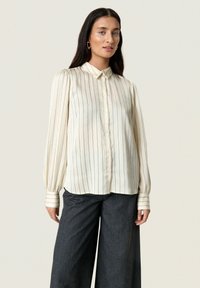Cream-striped blouse with a button-down design, long puffed sleeves, and contrasting striped cuffs, paired with dark wide-leg trousers.