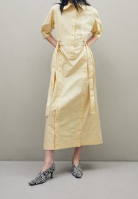 Kvinna klädd i en ljusgul knappad midiklänning med bälte och zebra-mönstrade högklackade skor, stående mot en enfärgad vägg.