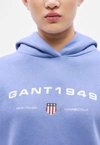 Sudadera con capucha azul claro con texto blanco que dice "GANT1949" y un emblema de escudo con estrellas y franjas. Tela suave, ajuste relajado.