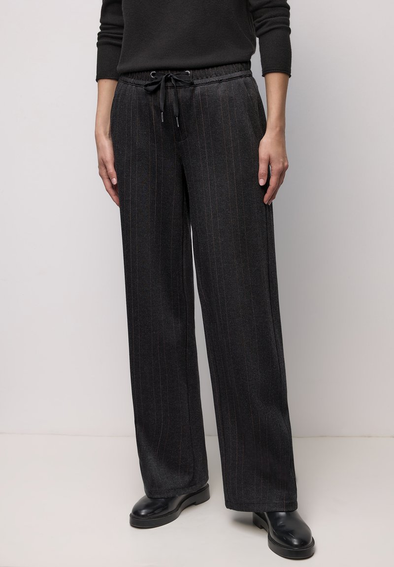 Vrouw draagt donkergrijze wijde pantalon met krijtstreep, een taille met trekkoord en zwarte leren enkellaarzen, voor een effen achtergrond.