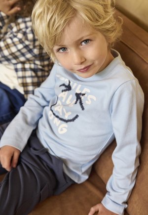Maglietta a maniche lunghe azzurro chiaro con testo bianco giocoso e grafica sorridente, abbinata a pantaloni scuri. Ragazzo seduto su un divano marrone.