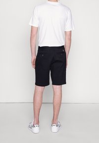 Sorte shorts med to baglommer, diskret stikninger og en gul logo-detail ved taljen. Kombineret med en hvid t-shirt og mønstrede sko.