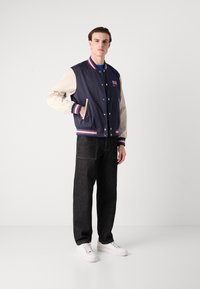 Marinblå och krämfärgad collegejacka med randiga mudd, tryckknappar och logotypdetalj, kombinerad med mörka jeans och vita sneakers.