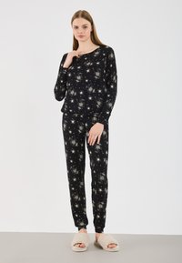 Ensemble de pyjama noir en tissu doux, affichant un motif d'éclat d'étoiles, avec des manches longues et un pantalon fuselé ; associé à des chaussons en peluche beiges.