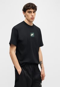 Νέος άνδρας με σγουρά μαλλιά, φορώντας μαύρο T-shirt Nike και μαύρο σορτς, στέκεται με το ένα χέρι στην τσέπη μπροστά σε απλό φόντο.