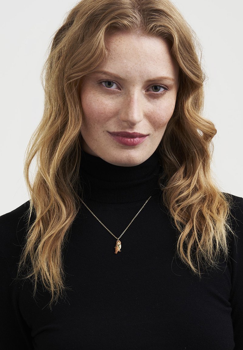 Frau mit welligem blondem Haar, die einen schwarzen Rollkragenpullover und eine goldene Halskette mit einem Eulen-Anhänger vor einem schlichten hellen Hintergrund trägt.