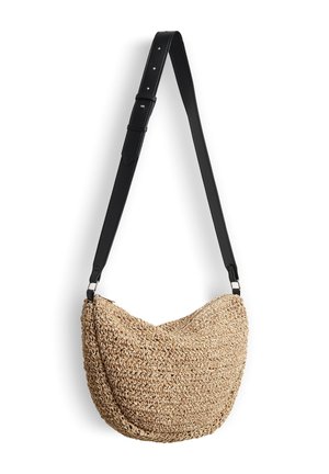 Geflochtene Strohtasche in geschwungener Form mit verstellbarem schwarzem Lederband, die vor weißem Hintergrund hängt.