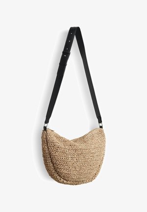 Geflochtene Strohtasche in geschwungener Form mit verstellbarem schwarzem Lederband, die vor weißem Hintergrund hängt.