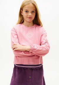 Jasnoróżowa bluzka z długim rękawem z haftowanym wzorem serca, zgromadzonymi mankietami i krótkim krojem, noszona na fioletowej spódnicy z korduroju.