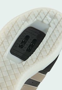 Podeszwa sneakersów z teksturowanym białym gumowym wykończeniem, czarnym wkładem z dwoma otworami na śruby oraz akcentami w kolorze brązowym i tan.
