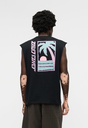 Persona con vitiligo de espaldas, vistiendo una camiseta negra sin mangas con un gráfico en tonos pastel de palmas y puesta de sol, junto con el texto "Santa Cruz".